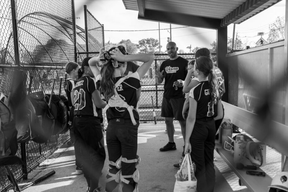 A youth softball coach crouches to talk with his team gathered in the dugout, players in full uniform and gear listening closely before taking the field.