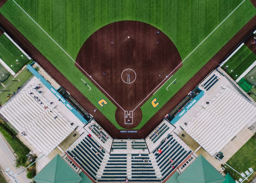 Aerial drone view straight down onto a college baseball/softball diamond and stadium seating, symmetric infield, foul lines, and dugouts at Frost Stadium.