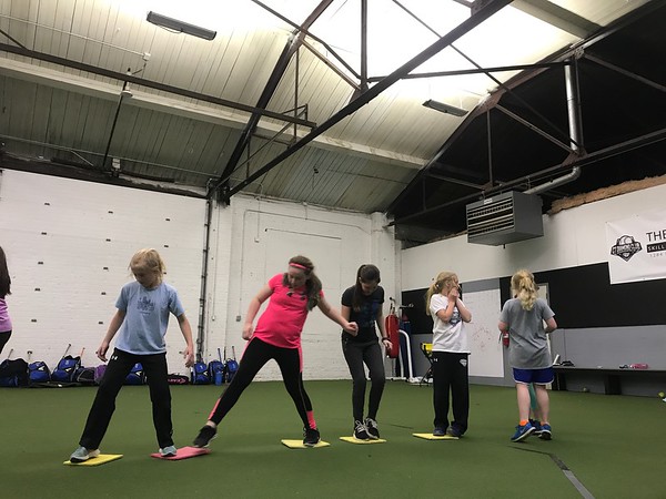 A Diamond Softball Club coach in a pink top leads five players through a stretching and mobility drill on the indoor turf, standing in among the group rather than off to the side.