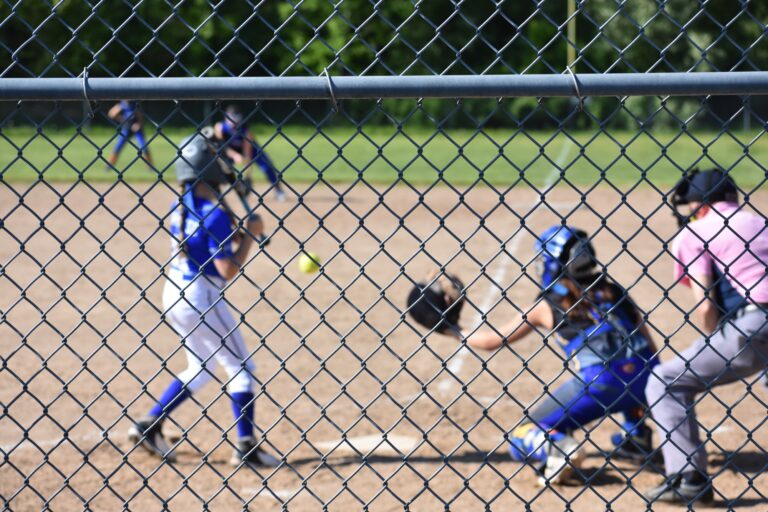 View through the backstop fence at a Diamond Softball Club game as a batter waits on the pitch and the catcher reaches for the ball